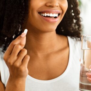 Smiling Afro woman holding a vitamin pill and a glass of water, symbolizing the beauty and wellness benefits of Howganics Biotin Shot